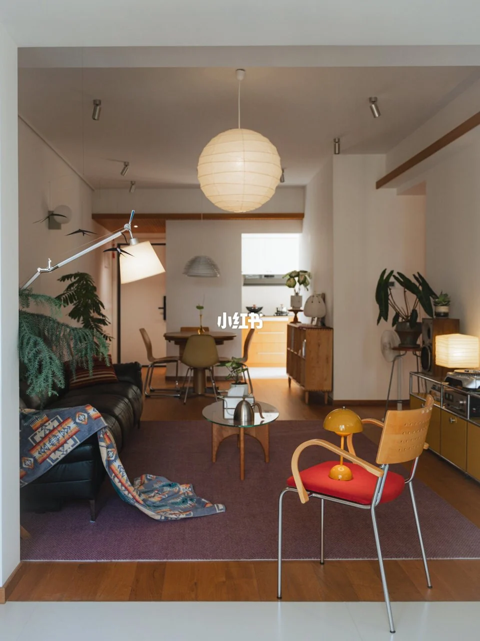 Black sofa, purple rug, yellow storage, red seated chair and paper pendant in a living room connected to dining.