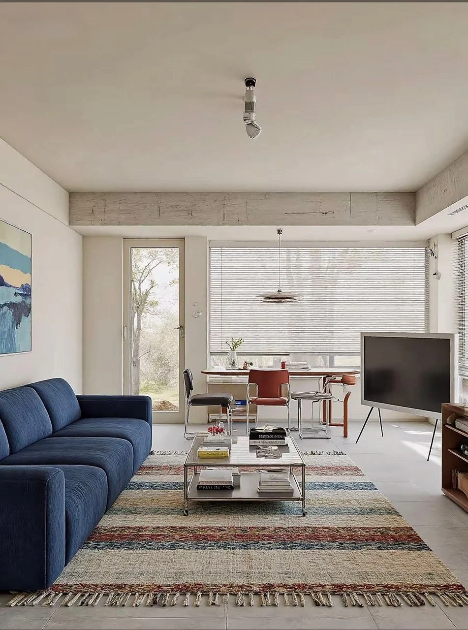 Deep blue sofa on a striped woven rug, glass coffee table and dining chairs beside a large window over gray tile.