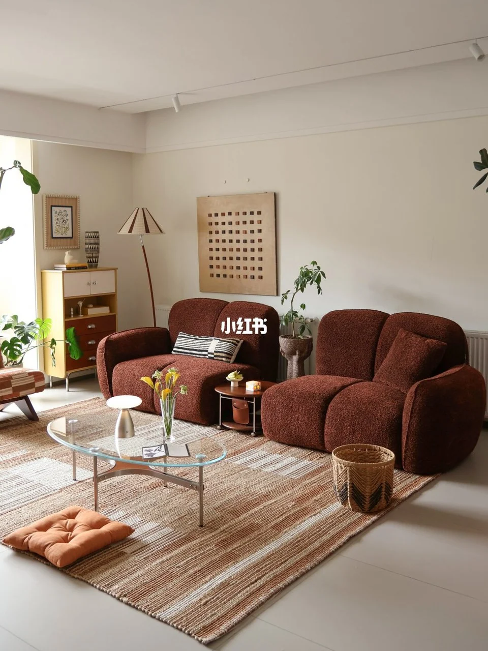 Textured brown seating, a tan striped rug, glass and chrome coffee table, plants and a pleated floor lamp over pale gray tile.