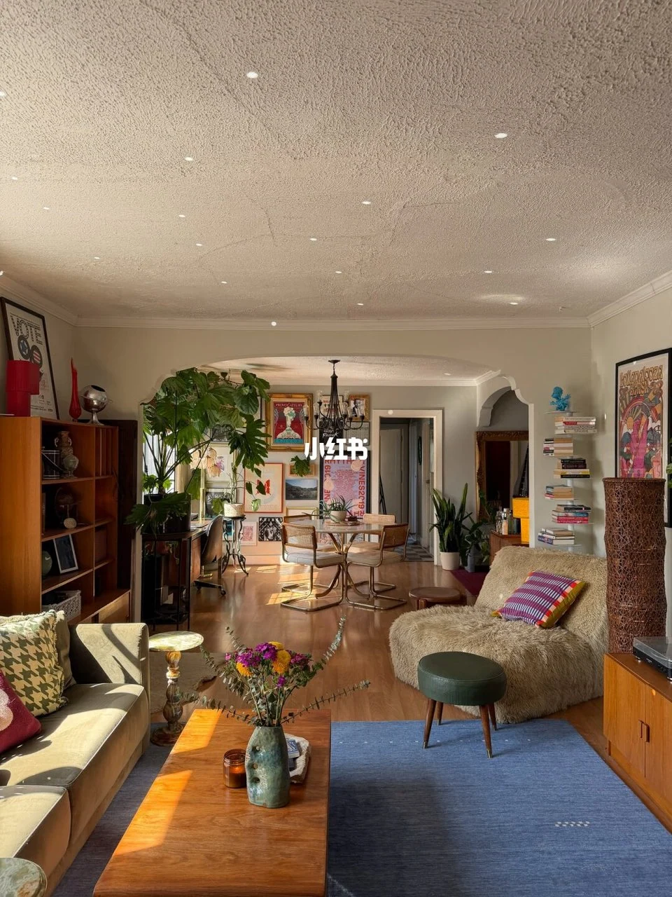Warm wood furniture, a blue rug, shaggy lounge chair and gallery wall in a connected living and dining room.