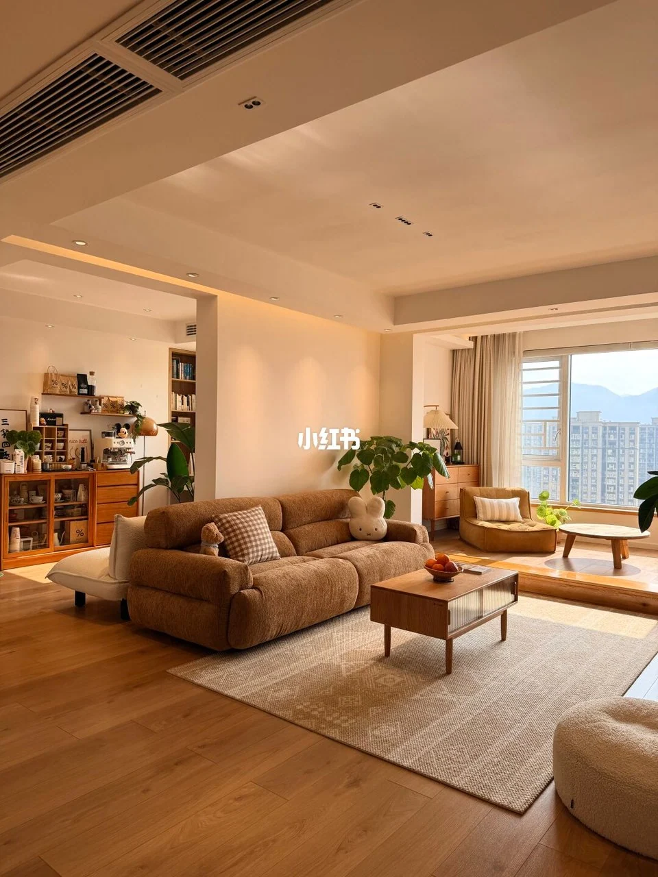 Soft brown sofa, pale rug and wood coffee table in a warm living room with a raised window sitting area and plants.