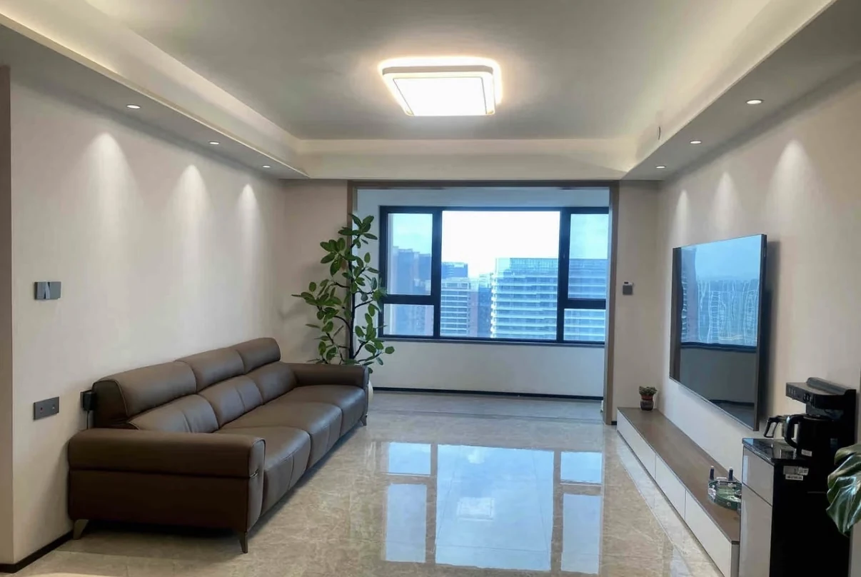 Original narrow living room with a brown leather sofa, reflective gray floor tile, cool blue-tinted window, television and coffee counter.