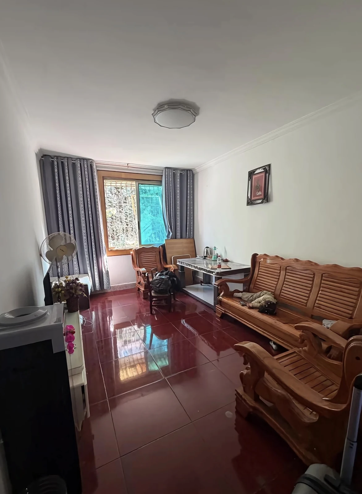 Original narrow living room with burgundy square tile, white walls, wooden seating and a wood-framed window.