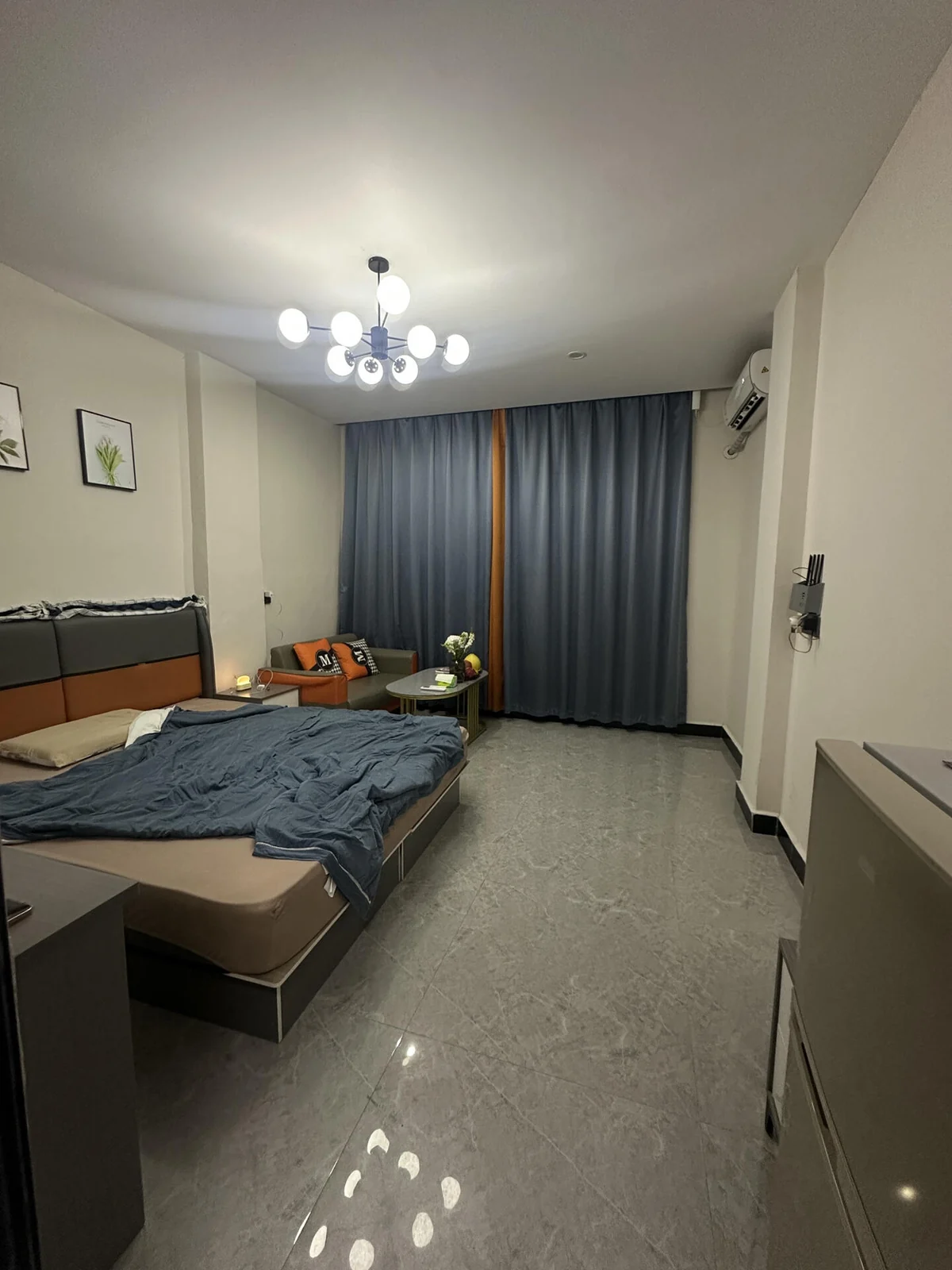Original small rental room with gray tile, a bed, compact sofa, oval coffee table, dark curtains and globe ceiling light.